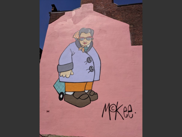 Large cartoon-style wall mural of an elderly woman in coat and sunshades pulling a shopping trolley bag